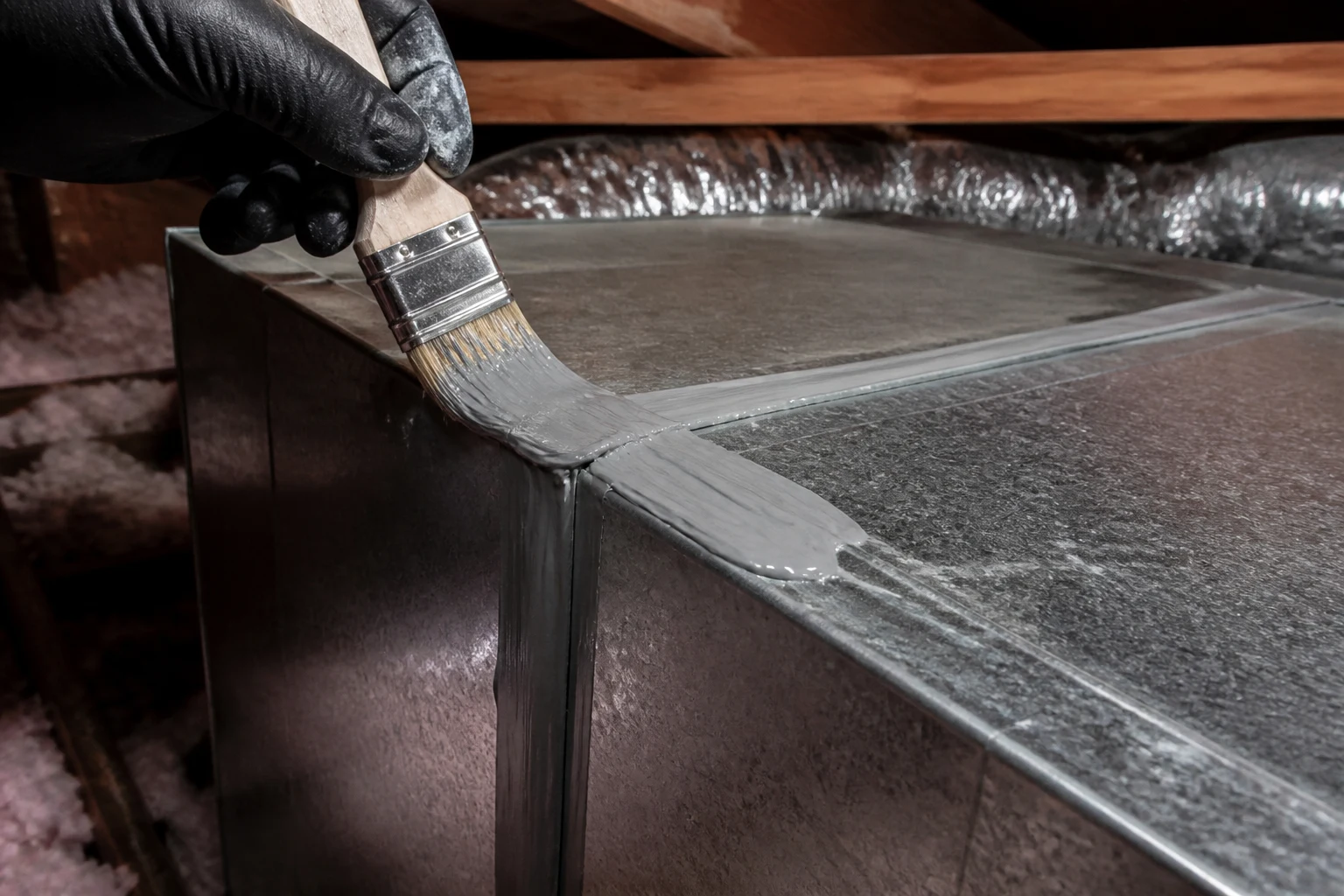 Teknisi HVAC sedang mengaplikasikan sealant mastik dengan kuas pada sambungan ducting logam di loteng